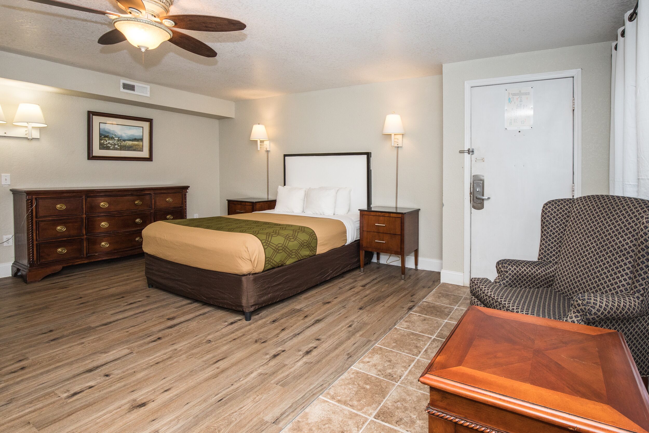 Habitación de hotel con cama, cómoda de madera, mesitas de noche, una silla y un ventilador de techo.