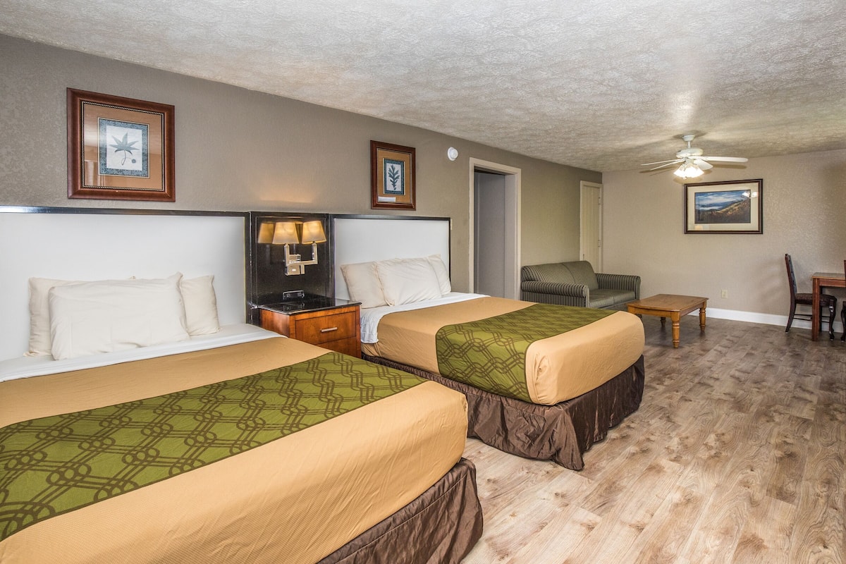 A hotel room with two beds, a nightstand with a lamp, a chair, a small table, and a ceiling fan.