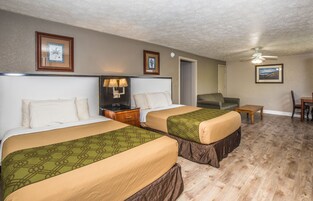 A hotel room with two beds, a nightstand with a lamp, a chair, a small table, and a ceiling fan.