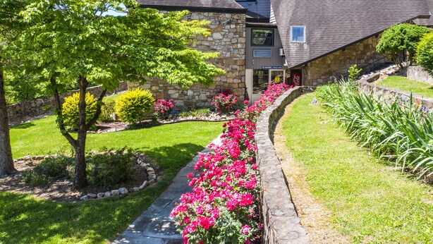 A stone building with a stone pathway lined with pink flowers, surrounded by green grass and trees. - Chalet Inn (Gatlinburg)