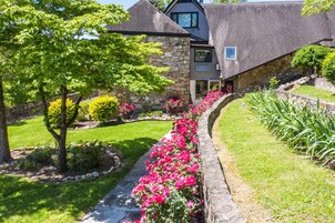 Un edificio de piedra con un camino empedrado bordeado de flores rosadas, rodeado de césped verde y árboles.