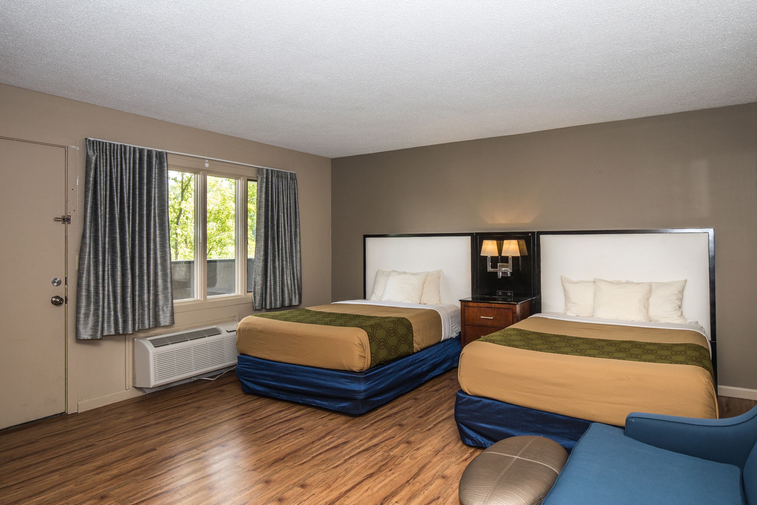 A hotel room with two beds, a window with curtains, a heater, and a small table with a lamp.