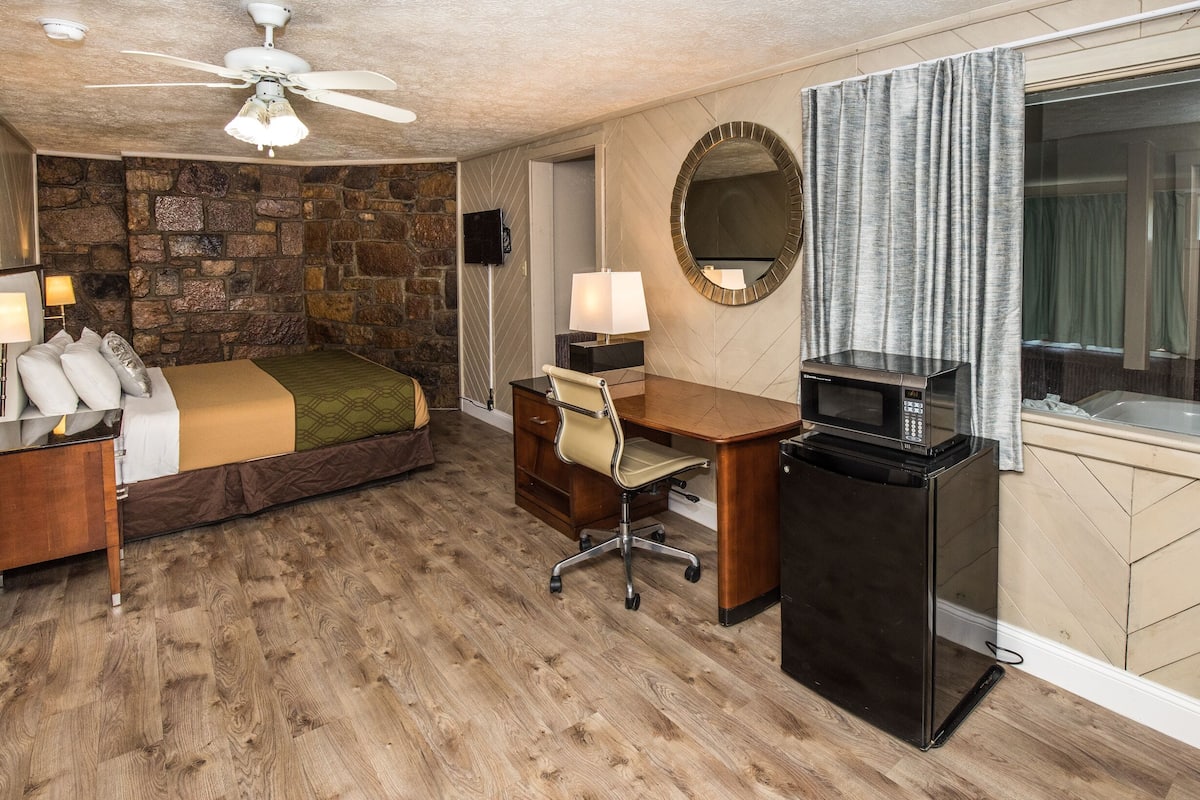 A hotel room with a stone wall, a bed with a green blanket, a desk with a lamp, a TV, and a microwave.