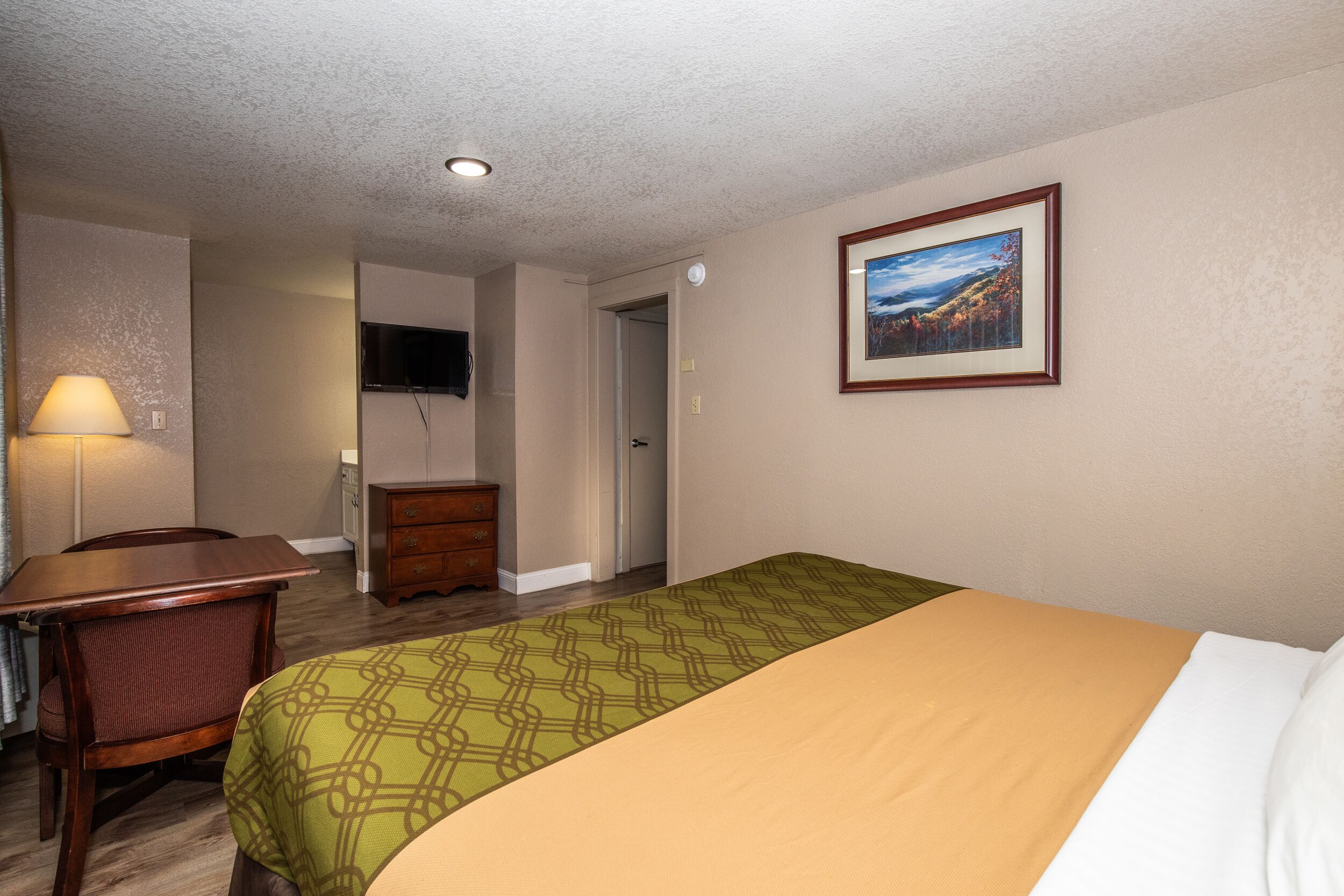 A hotel room with a bed, a desk, a chair, a dresser, and a television on the wall.