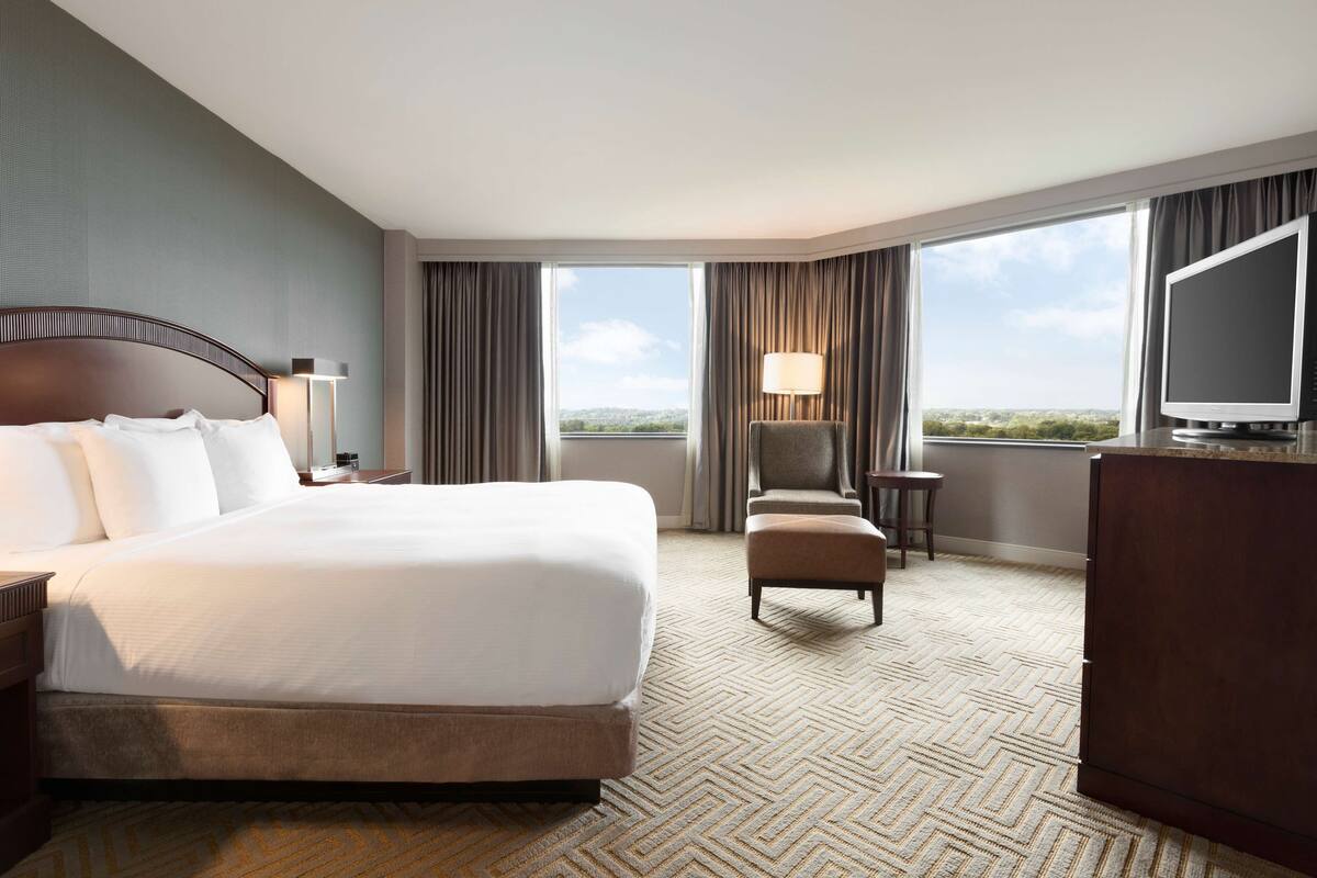 A hotel room with a large bed, a desk, a chair, a TV, and a view of the city through the windows.
