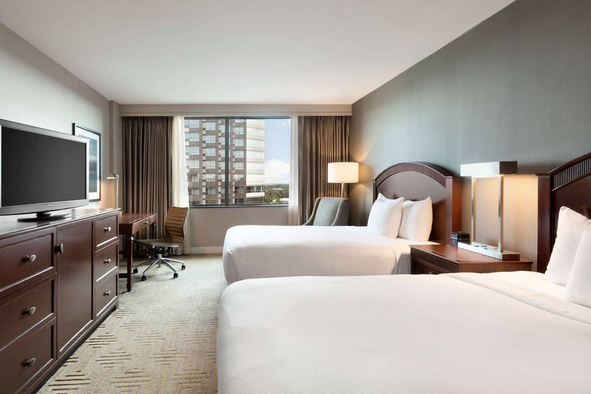 Ein Hotelzimmer mit zwei Betten, einem Fernseher, einem Schreibtisch mit Stuhl und einem großen Fenster mit Blick auf ein Gebäude.