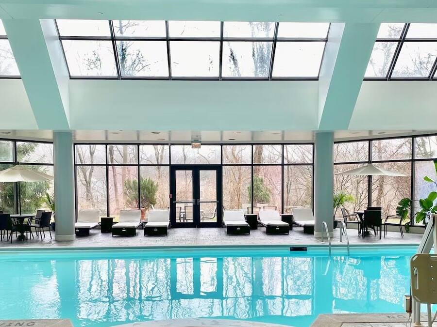 An indoor swimming pool with clear glass ceilings, lounge chairs, and potted plants.