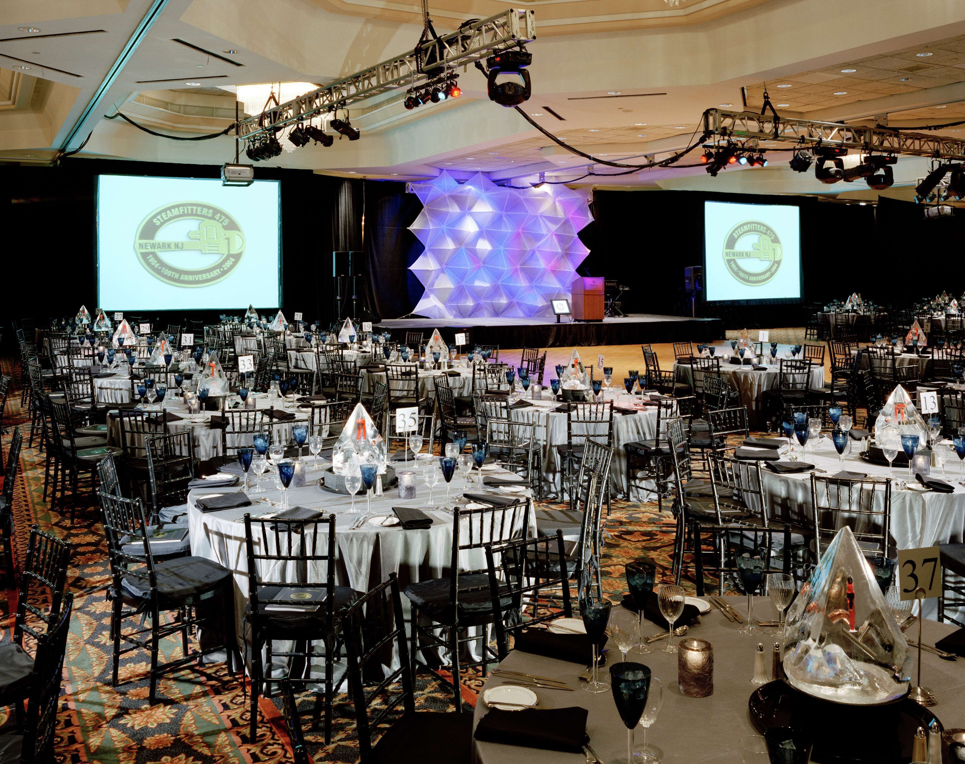 A banquet hall with round tables, chairs, and a stage with a geometric light installation.