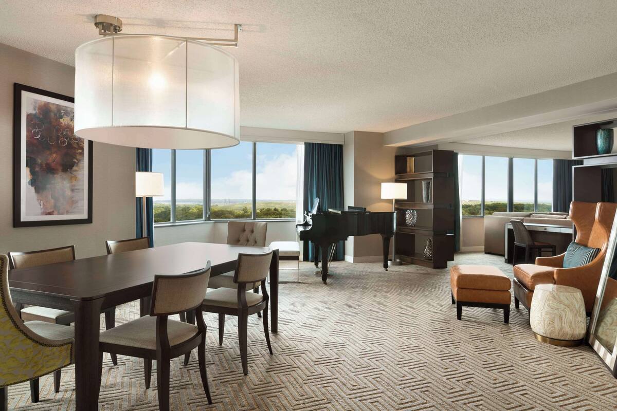A spacious hotel room with a dining area, a piano, and a large window offering a view of the landscape.