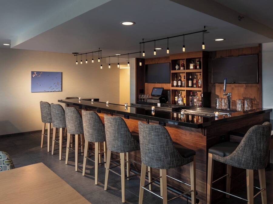 Een bar met een lange toonbank, verschillende krukken, een aan de muur bevestigde televisie en een plank met flessen.