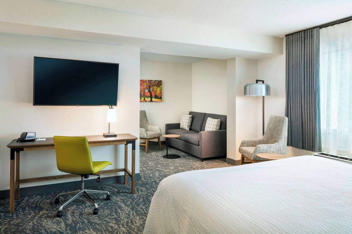 A hotel room with a bed, a desk with a chair, a sofa, a TV, and a lamp.