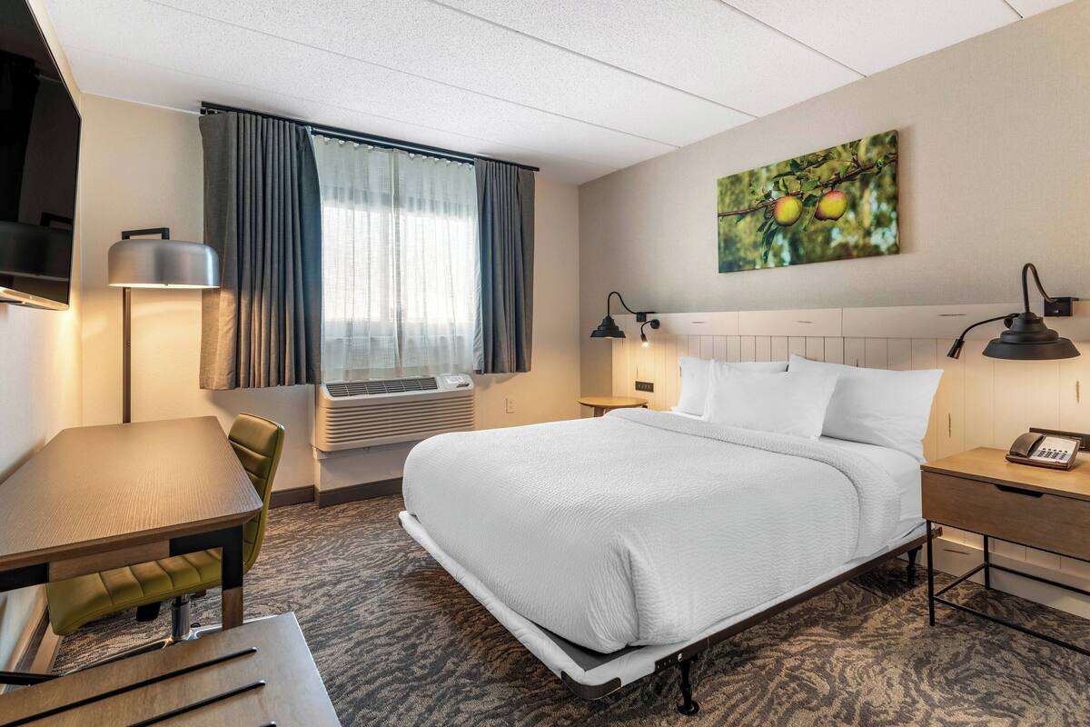 A hotel room with a bed, a desk, a chair, a lamp, a painting, and an air conditioning unit.