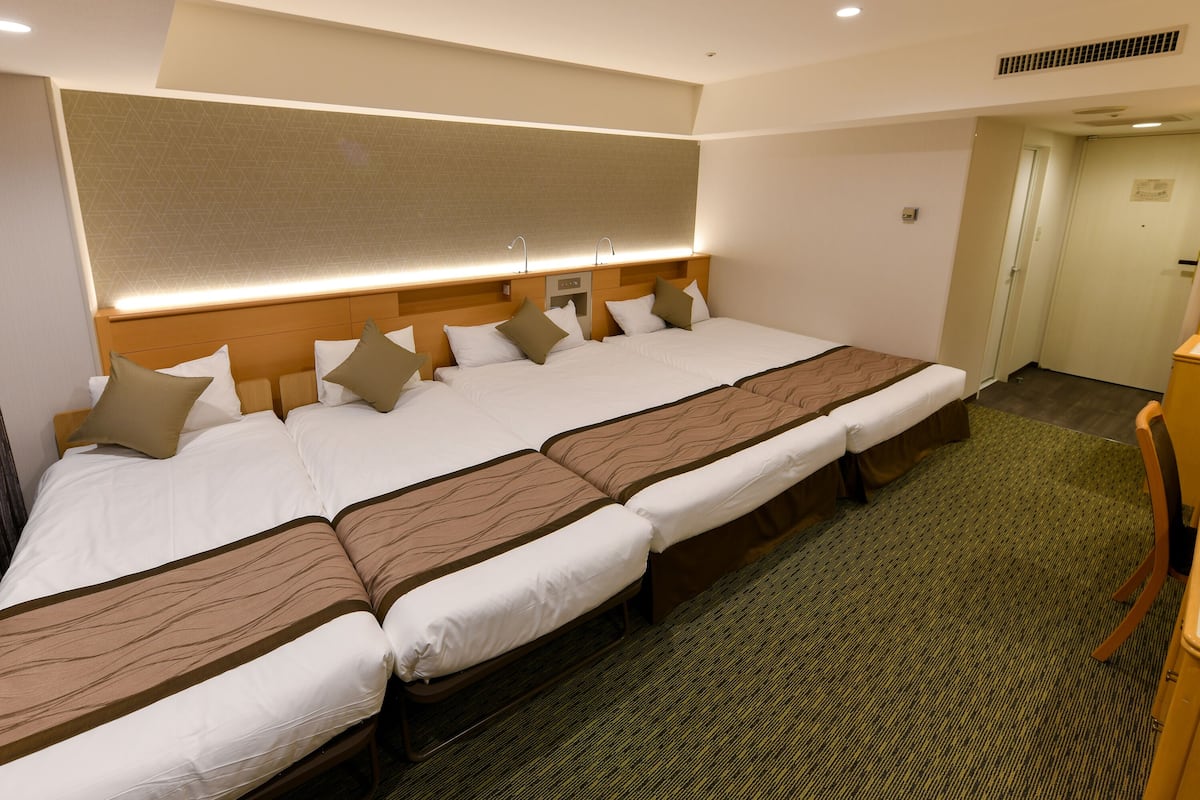 A hotel room with a large bed, a headboard with built-in lighting, a carpeted floor, and a door leading to another room.