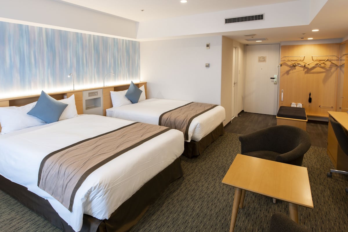 A hotel room with two beds, a desk, a chair, and a coat rack.