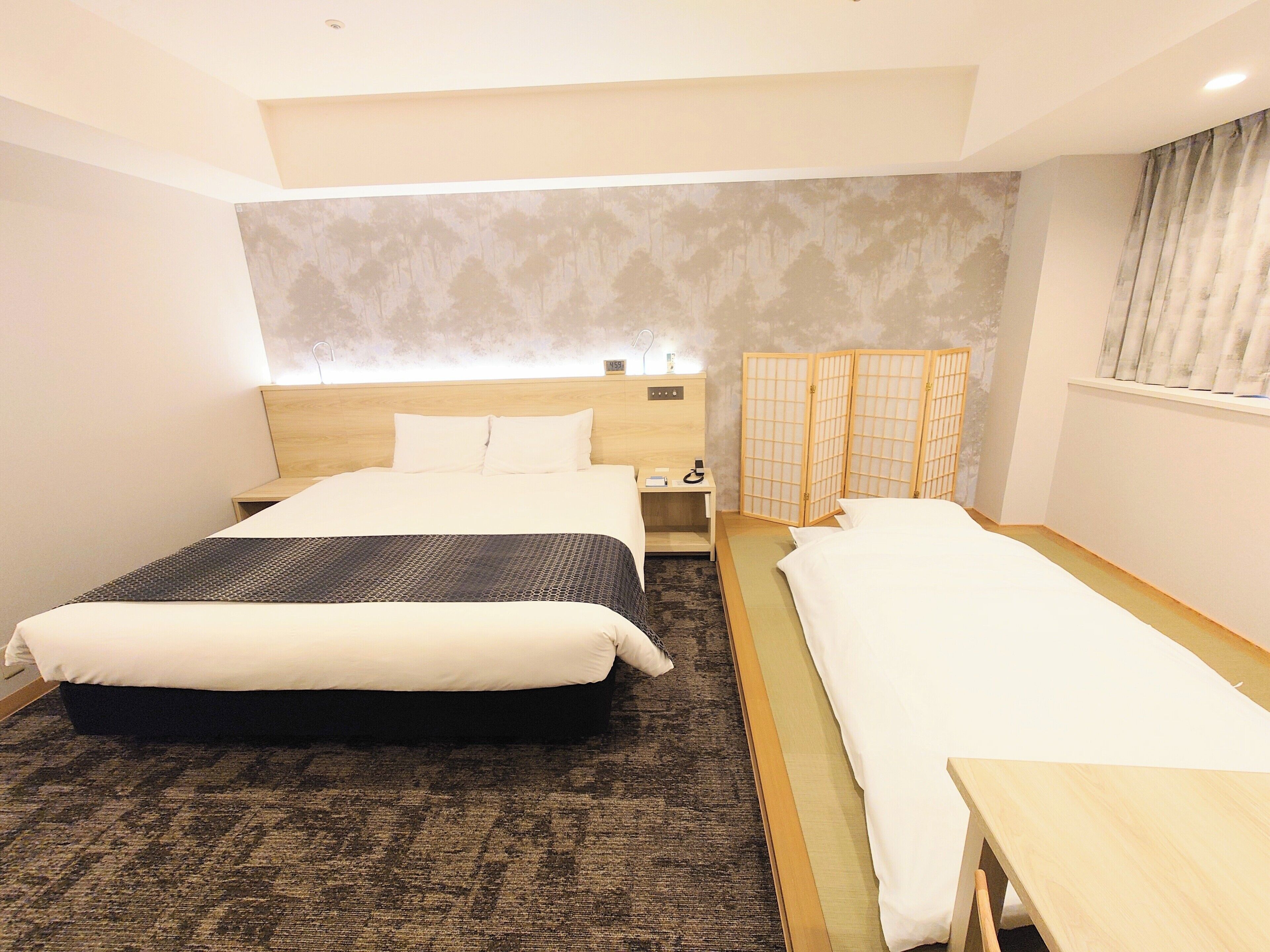 A hotel room with a large bed, a smaller bed, a wooden nightstand, a bedside table, a wall-mounted shelf, and a traditional Japanese room divider.
