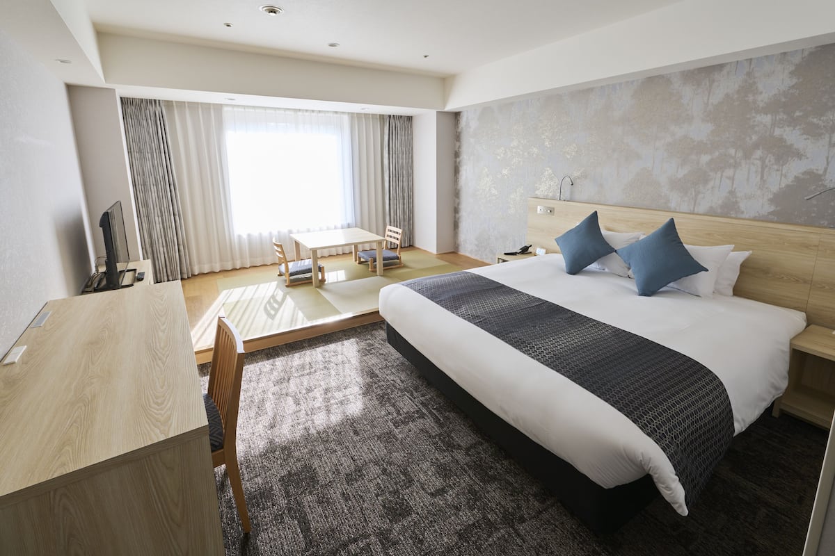 A hotel room with a large bed, a desk, and a chair. There is a window with curtains, a wall with a tree mural, and a nightstand with a lamp.