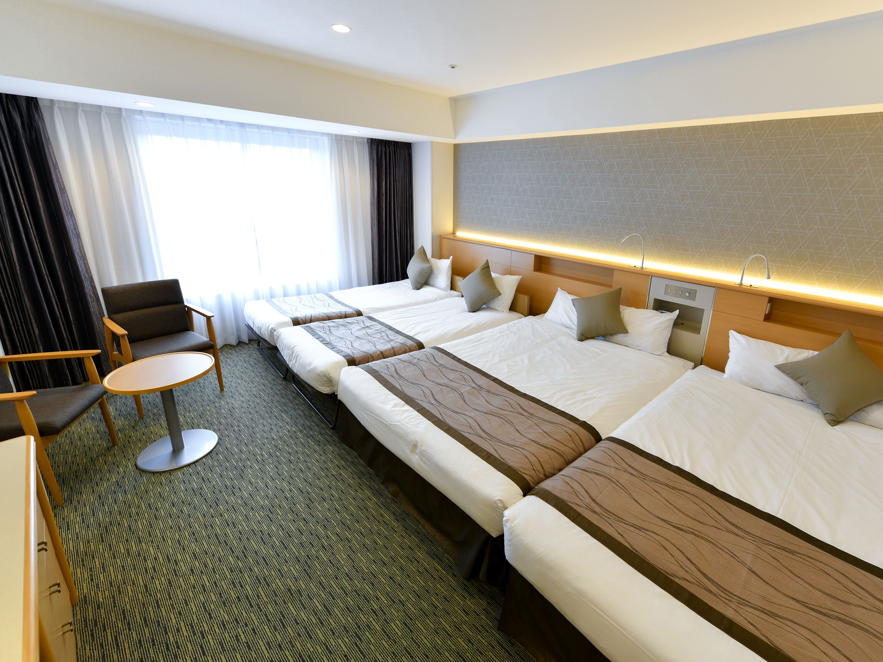 Habitación de hotel con dos camas individuales, un sofá, una mesita redonda y un ventanal con cortinas.