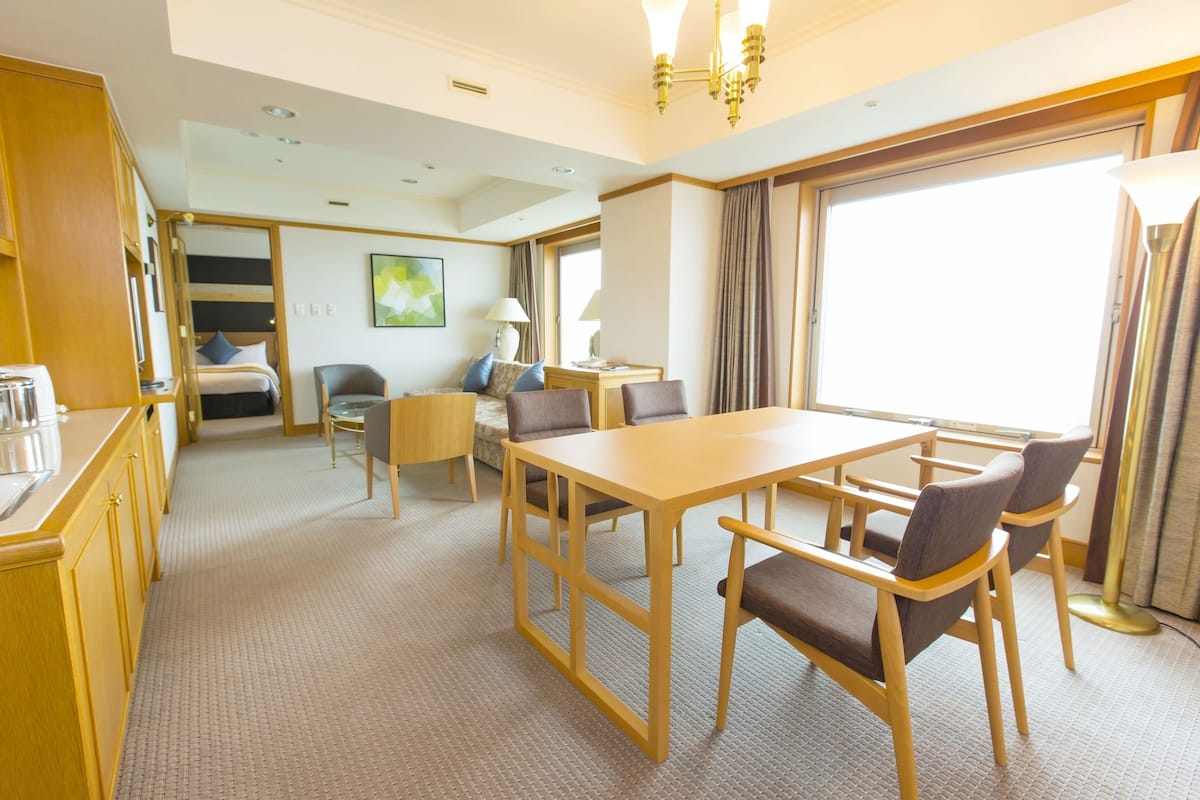 A hotel room with a wooden dining table, four chairs, a bed with a headboard, a desk with a chair, a lamp, and a large window with curtains.