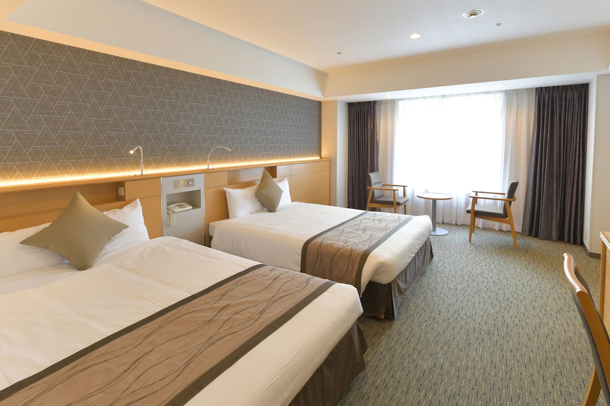 A hotel room with two beds, a small desk with a phone, a chair, a round table, and a large window with curtains.