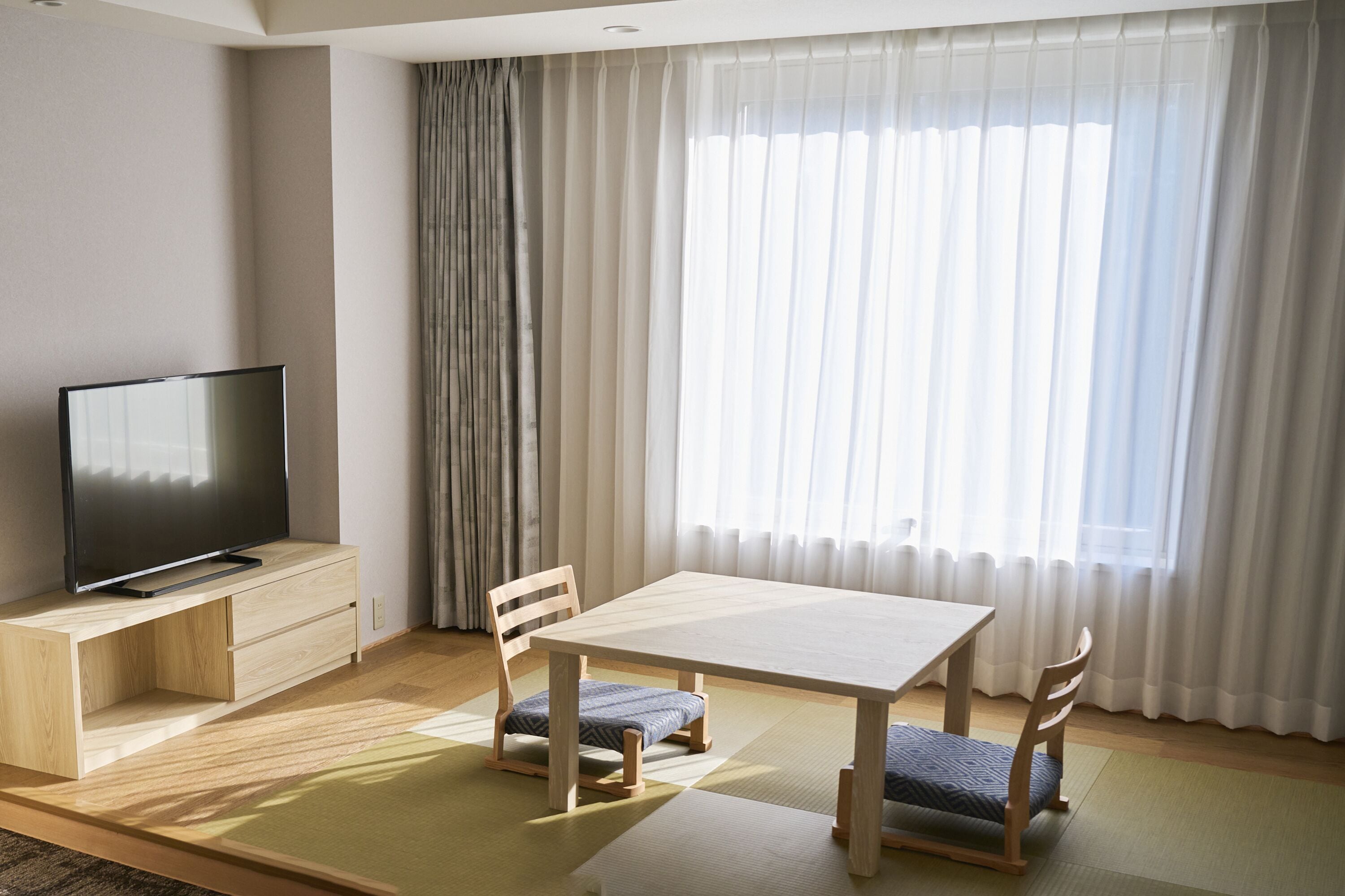 Un salon moderne avec un téléviseur à écran plat posé sur un meuble en bois, une table à manger en bois avec deux chaises, et une grande fenêtre ornée de rideaux légers.