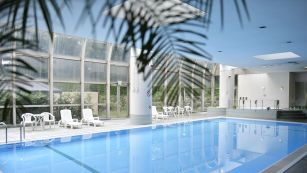 Una piscina con agua cristalina azul, rodeada de sillas reclinables blancas y un muro de vidrio con ventanales amplios.