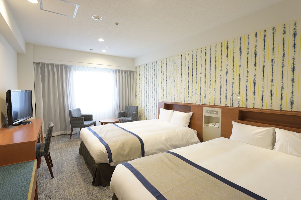 A hotel room with two beds, a desk with a television, a chair, and a window with curtains.