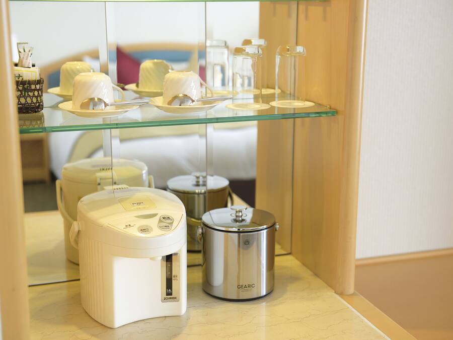 Una zona de cocina con cafetera, cocedor de arroz y repisa de vidrio que exhibe objetos de cerámica blanca.