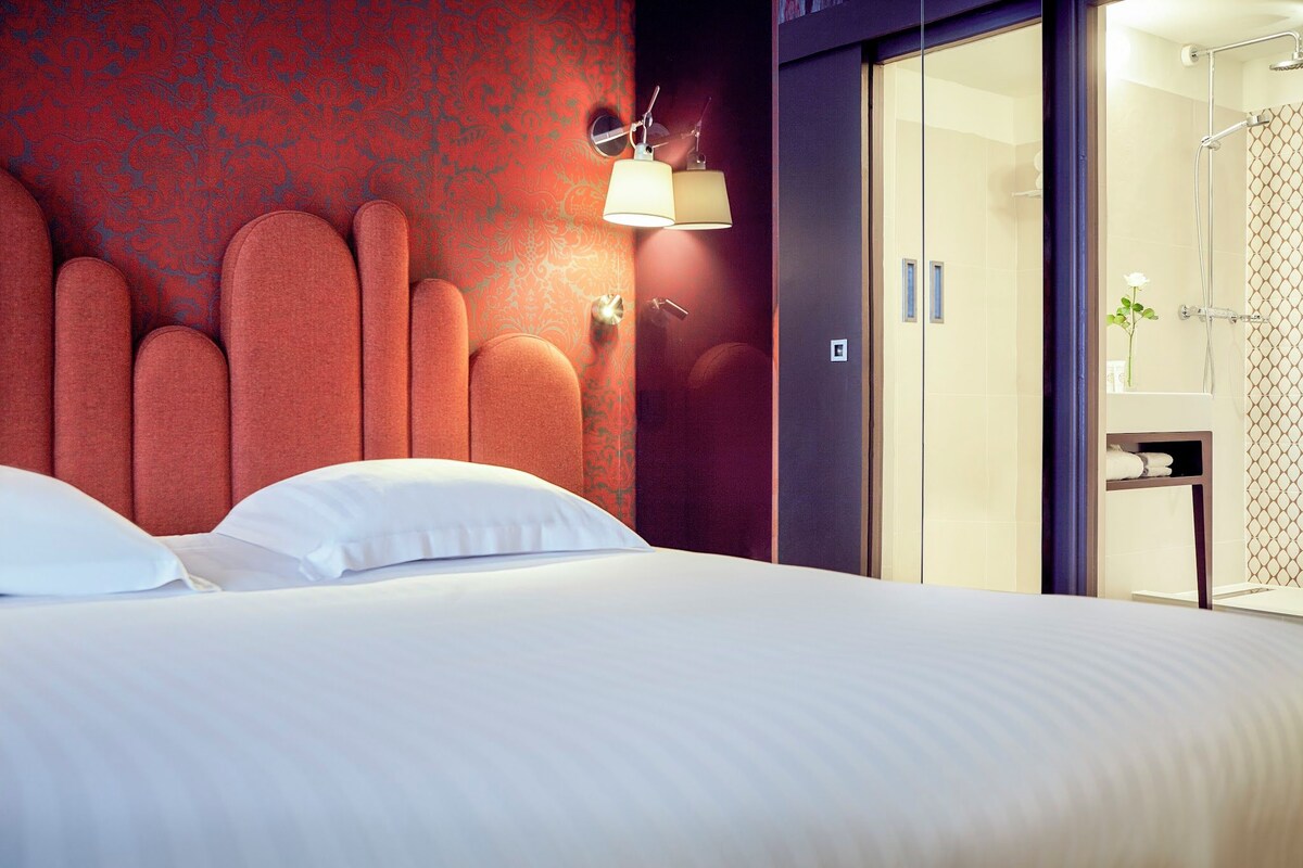A hotel room with a large bed, a red patterned wall, and a modern bathroom with a shower and a mirror.