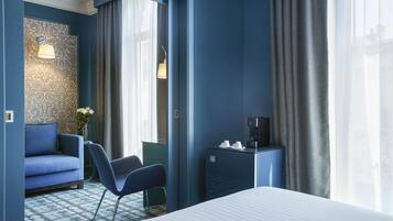 Une chambre d’hôtel avec un lit, un canapé bleu, une chaise, une petite table avec une machine à café et un mur recouvert d’un papier peint à motifs.