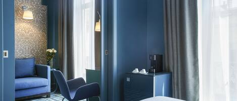 A hotel room with a bed, a blue sofa, a chair, a small table with a coffee maker, and a wall with a patterned wallpaper.