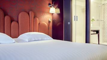 Une chambre d’hôtel dotée d’un grand lit, d’une tête de lit au design original, d’une lampe fixée au mur et d’une salle de bain ouverte avec douche.