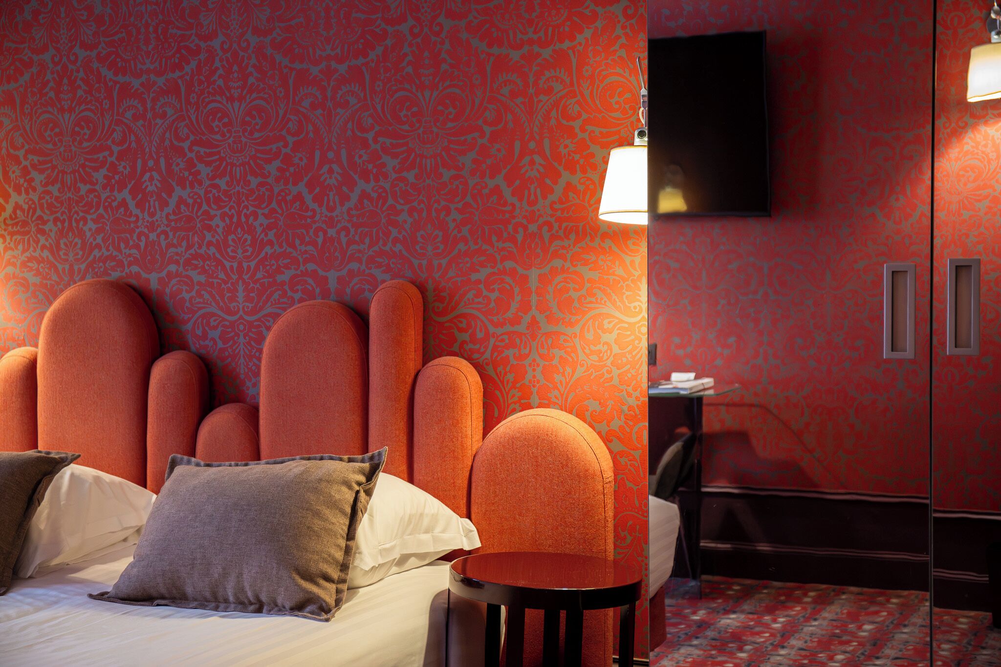 A hotel room with a patterned wallpaper, a bed with pillows, a chair, a small table, and a TV mounted on the wall.
