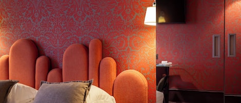 A hotel room with a patterned wallpaper, a bed with pillows, a chair, a small table, and a TV mounted on the wall.