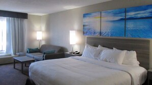 A hotel room with a large bed, two bedside tables, a chair, a sofa, and a painting of a sunset over water on the wall. - Clarion Pointe Evansville North (Evansville)