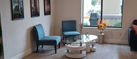 A modern living room with a glass coffee table, two blue armchairs, and a large window with a view of the outdoors.