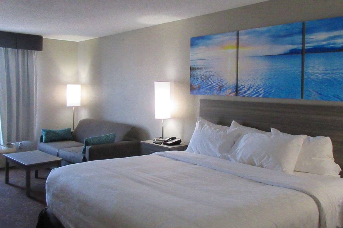 A hotel room with a large bed, a sofa, a chair, a small table, and a nightstand with a lamp.