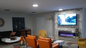 A modern living room with a fireplace, a flat-screen TV mounted on the wall, and a seating area with orange chairs. - Clarion Pointe Evansville North (Evansville)