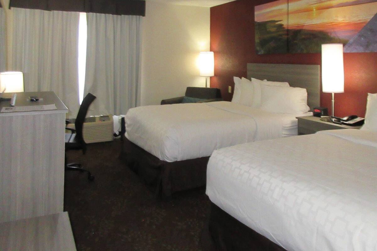 A hotel room with two beds, a desk, a chair, a lamp, and a painting on the wall.