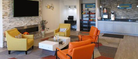 A living room with yellow and orange chairs, a flat-screen TV, a fireplace, and a kitchen area with a stone backsplash and a sign that reads "NOURISH".