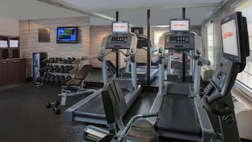 Academia com esteiras, halteres e uma televisão exibindo programas de exercícios.