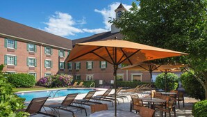A poolside area with lounge chairs and tables, a brick building in the background, and a clear blue sky. - Sonesta Select Boston Lowell Chelmsford  (Lowell)