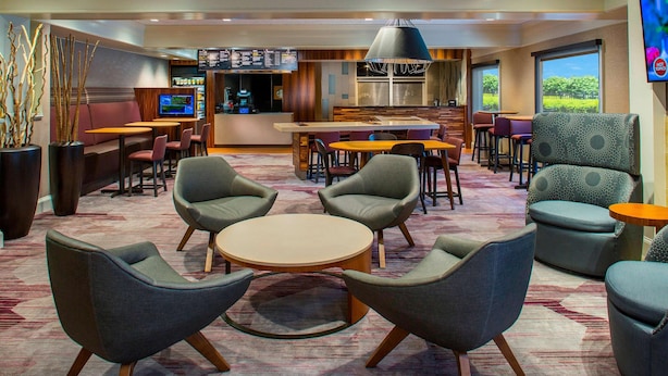 A modern hotel lobby with a central seating area, a bar, and a large window offering a view of the outdoors. - Sonesta Select Boston Lowell Chelmsford  (Lowell)