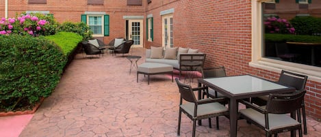 A courtyard with a brick building, green shutters, a seating area with a table and chairs, and a hedge with pink flowers.