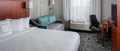 A hotel room with a large bed, a sofa, a desk with a TV, and an air conditioning unit.