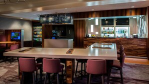A bar area with a long counter, multiple bar stools, a digital menu display, a refrigerator stocked with beverages, and a bar stocked with various bottles. - Sonesta Select Boston Lowell Chelmsford  (Lowell)