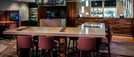 A bar area with a long counter, multiple bar stools, a digital menu display, a refrigerator stocked with beverages, and a bar stocked with various bottles.