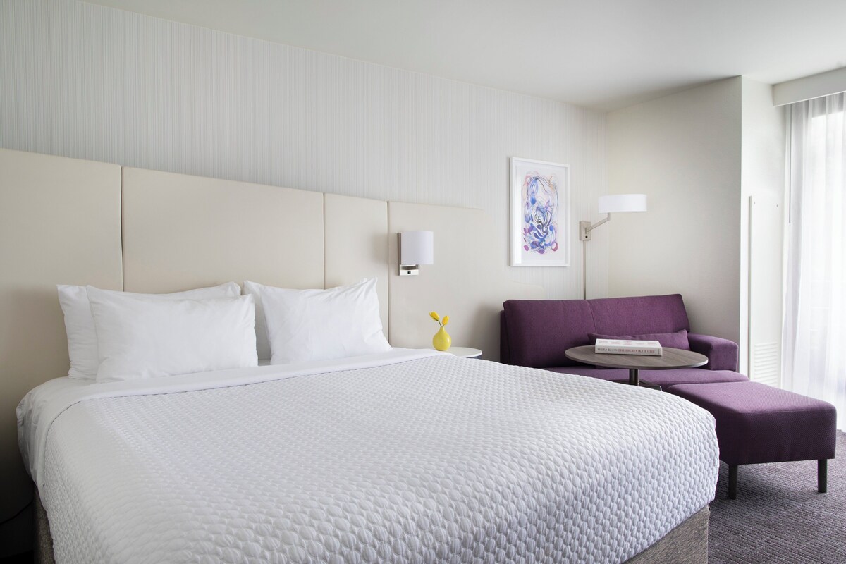 A hotel room with a large bed, a small sofa, a round table, and a wall-mounted lamp.
