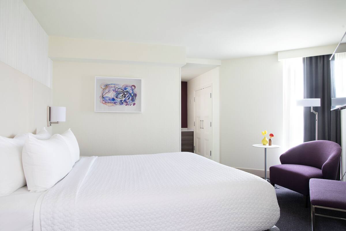 Un cuarto de hotel con una cama grande, un sillón morado, una pequeña mesa con flores y un cuadro en la pared.