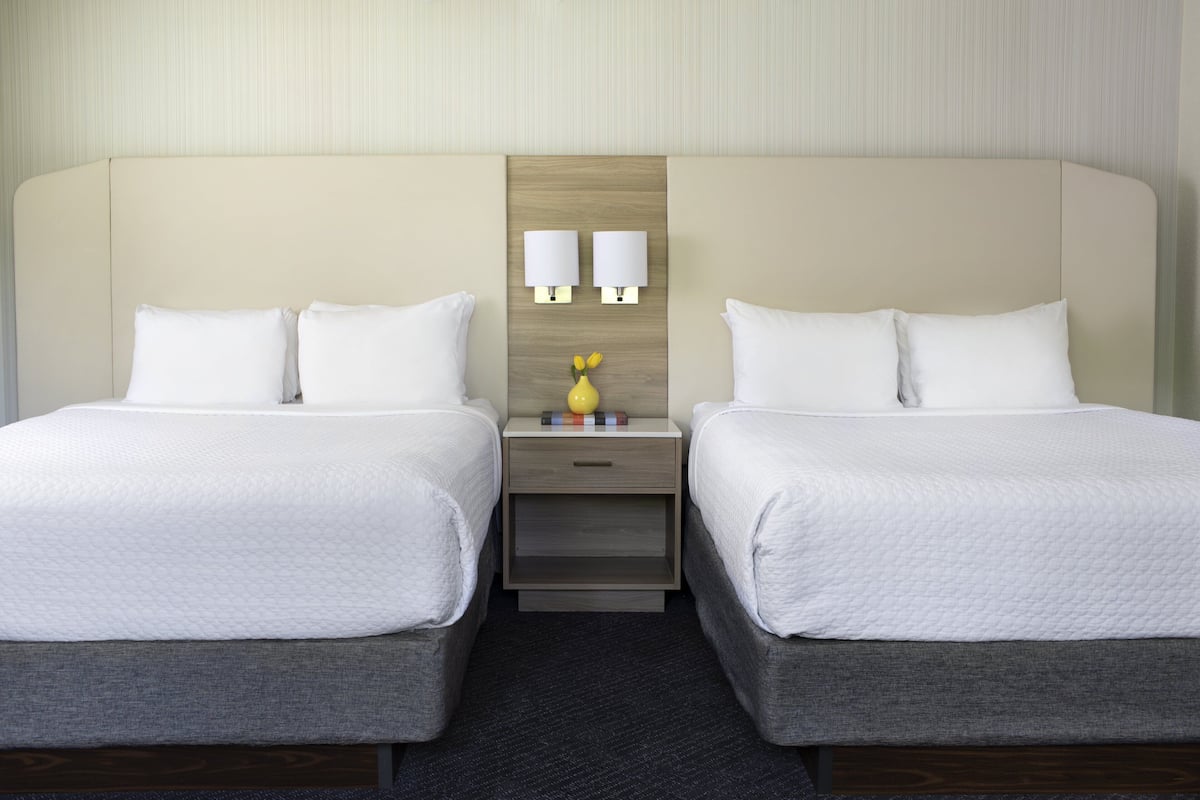 Habitación de hotel con dos camas individuales, una mesita de noche con dos lámparas fijadas a la pared y un pequeño florero con flores.