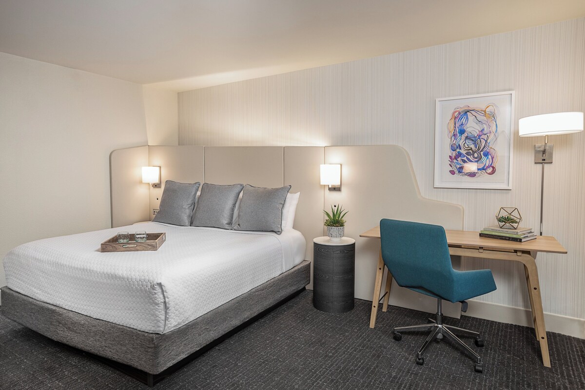 A hotel room with a bed, bedside table, lamp, desk, chair, and a framed artwork on the wall.